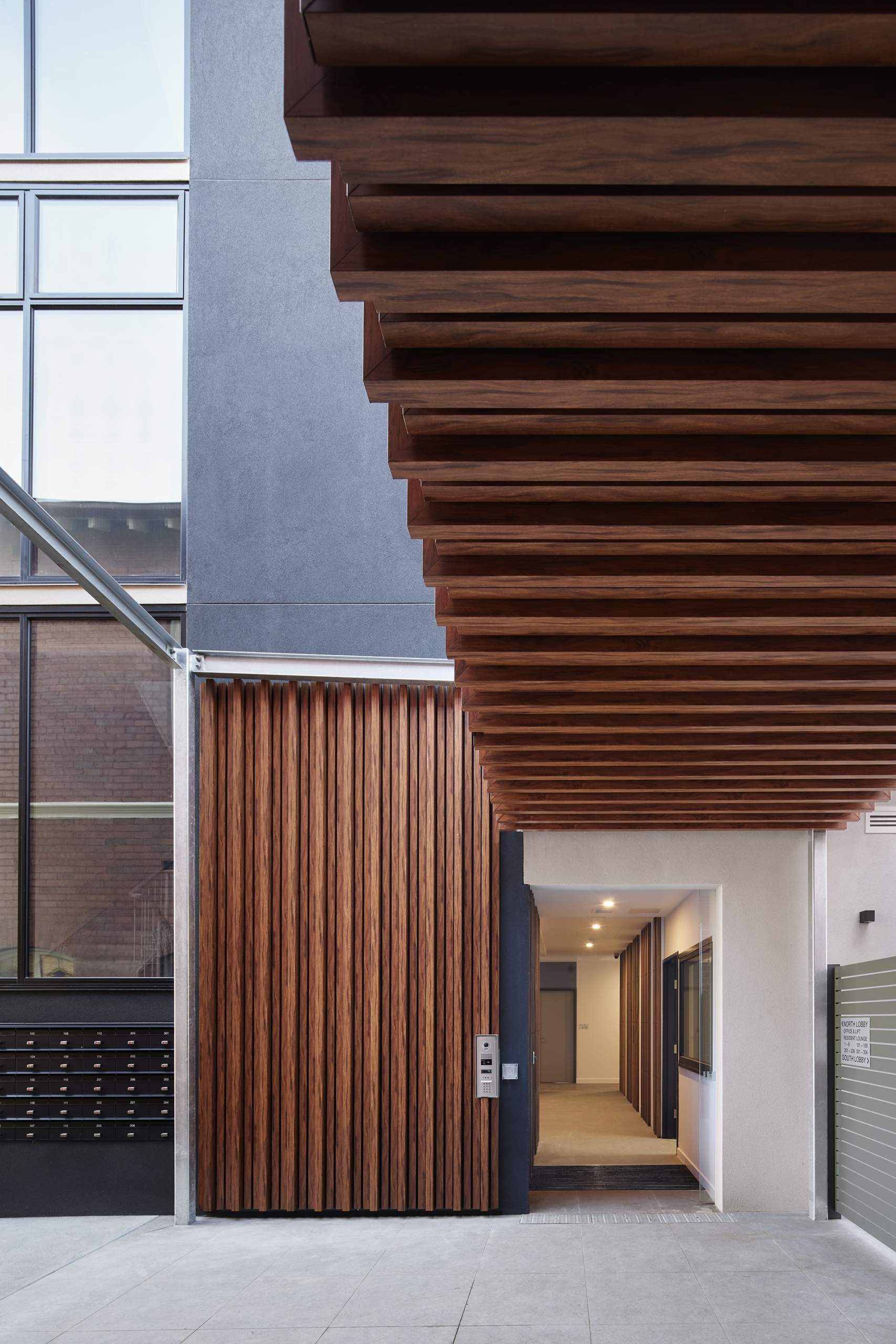 A modern building entrance with vertical wooden slats and large horizontal wooden beams overhead, leading to a well-lit hallway. Black mailboxes are mounted on the left wall.
