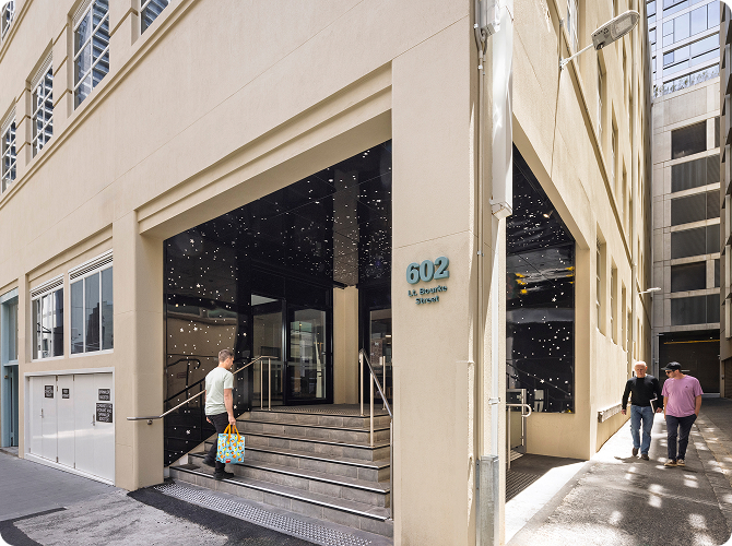 A man with a colorful bag walks up the steps to the entrance of a modern beige building at 602 Little Bourke Street, while two people walk by on the sidewalk.