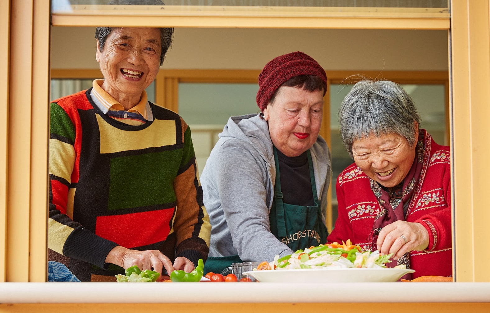 Three older adults stand together at a kitchen window, smiling and preparing a salad. They are dressed in colorful sweaters and appear to be enjoying each other’s company while cooking.