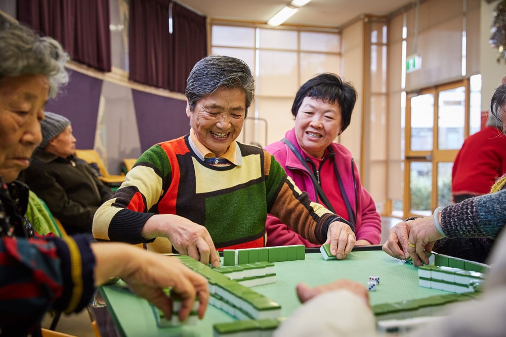 A group of elderly people sit around a table playing mahjong. They are smiling and appear to be enjoying each others company in a bright, indoor setting.