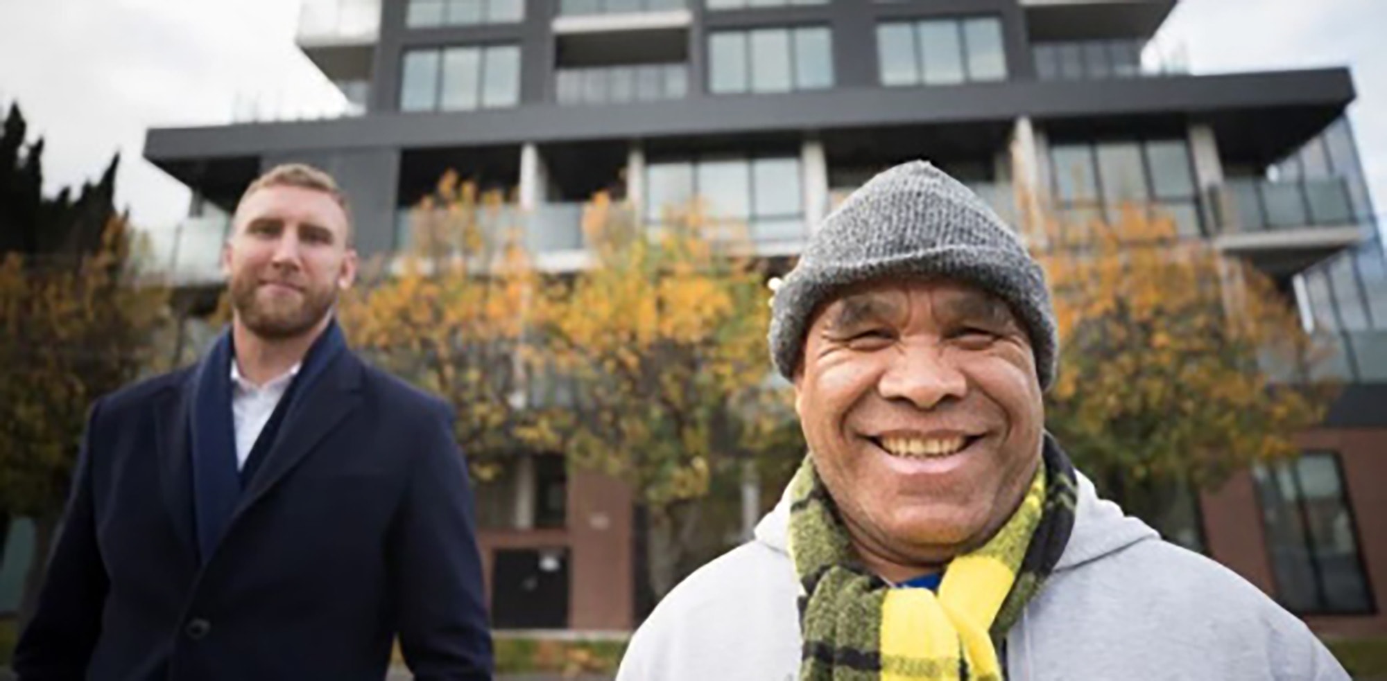 Two men stand outside in front of a modern apartment building. The man in the foreground is smiling, wearing a gray beanie and scarf. The man in the background is wearing a dark coat. Autumn trees are visible behind them.