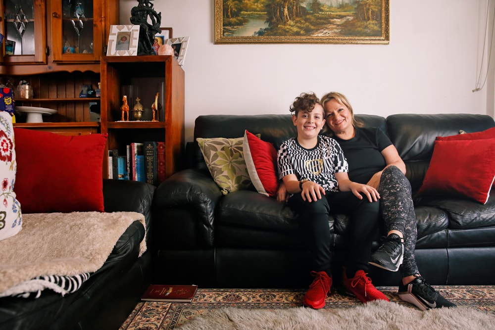A smiling woman and child sit together on a black couch in a cozy living room with red cushions, a bookshelf, and a painting on the wall behind them.