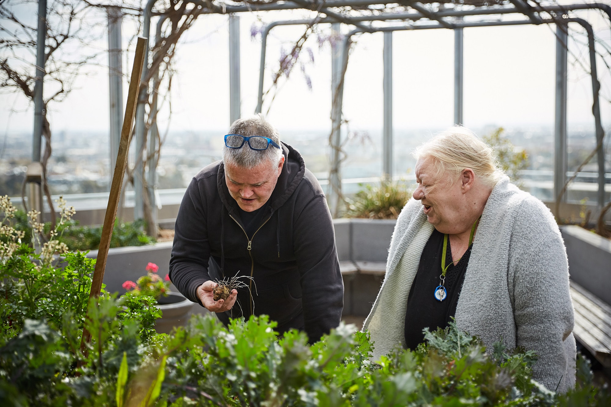 Two older adults are tending to plants in an outdoor rooftop garden. One person holds a small plant, while the other smiles and observes. The background shows trellises and a city view.