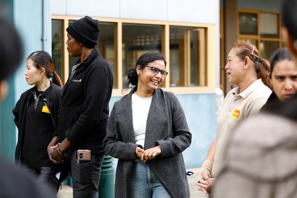 A group of people stands outside a building, casually talking and smiling. One woman in the center wearing glasses and a gray sweater laughs while interacting with others. The setting appears to be friendly and informal.