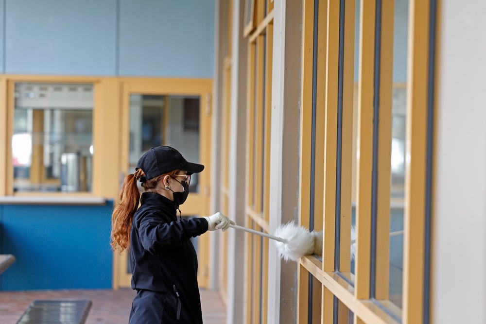 A person wearing a black cap, jacket, gloves, and mask uses a duster to clean large glass windows in a modern building with yellow window frames.