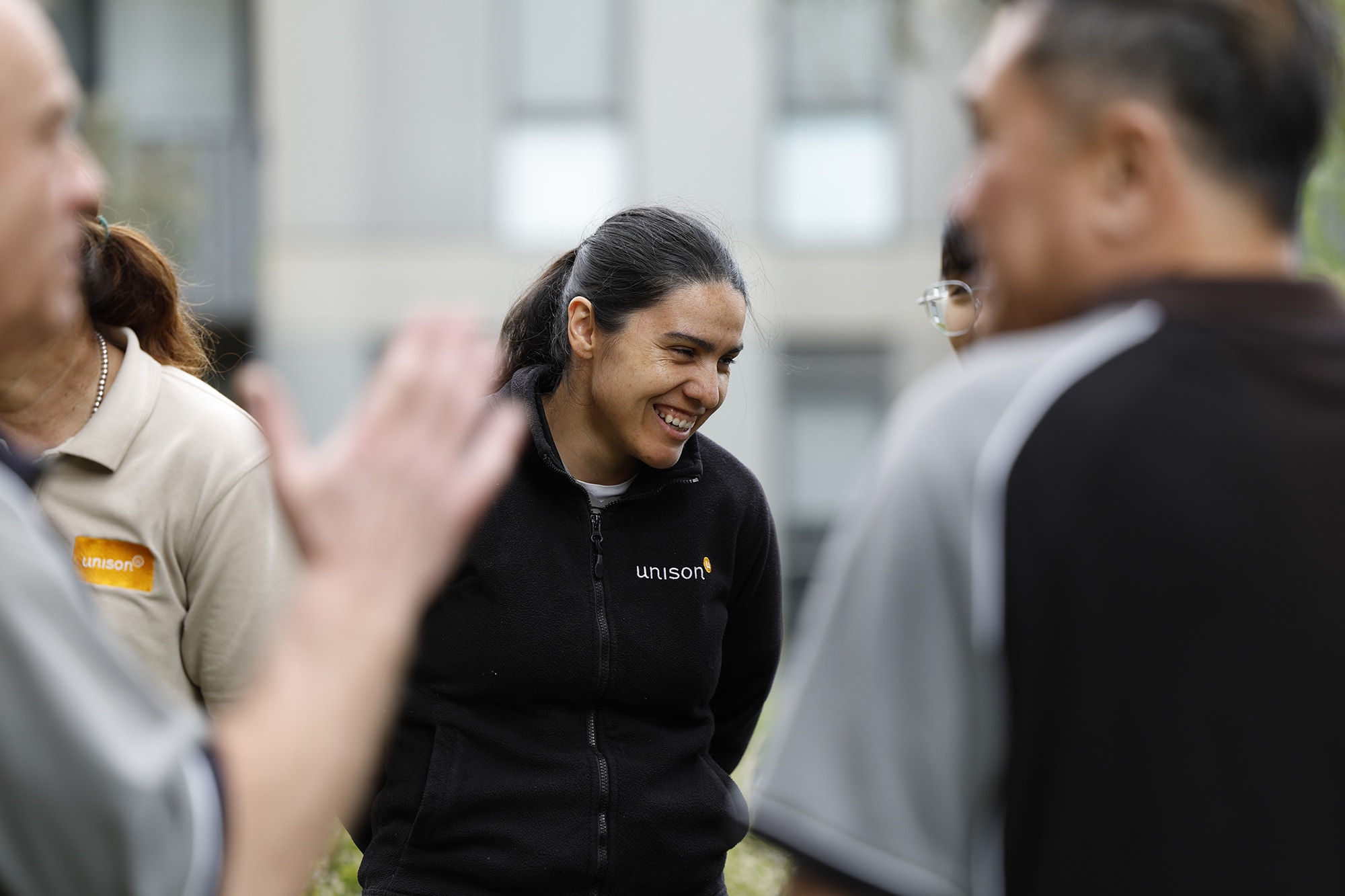 A woman in a black unison jacket smiles while standing outdoors with a group of people wearing uniforms. The background is blurred, focusing on the group interaction.