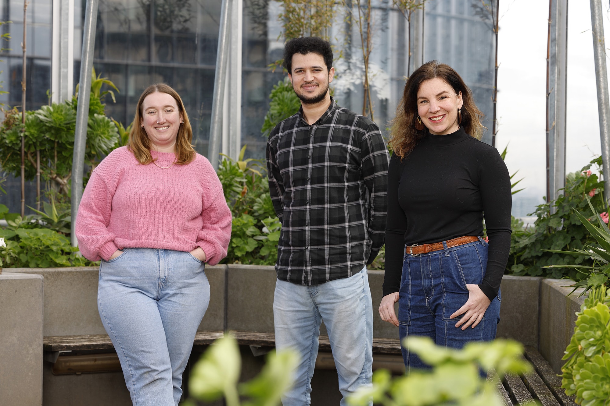 Three people stand outdoors in front of plants and modern glass buildings. They are smiling, with two women on either side wearing casual sweaters and jeans, and a man in the middle wearing a black checkered shirt and jeans.