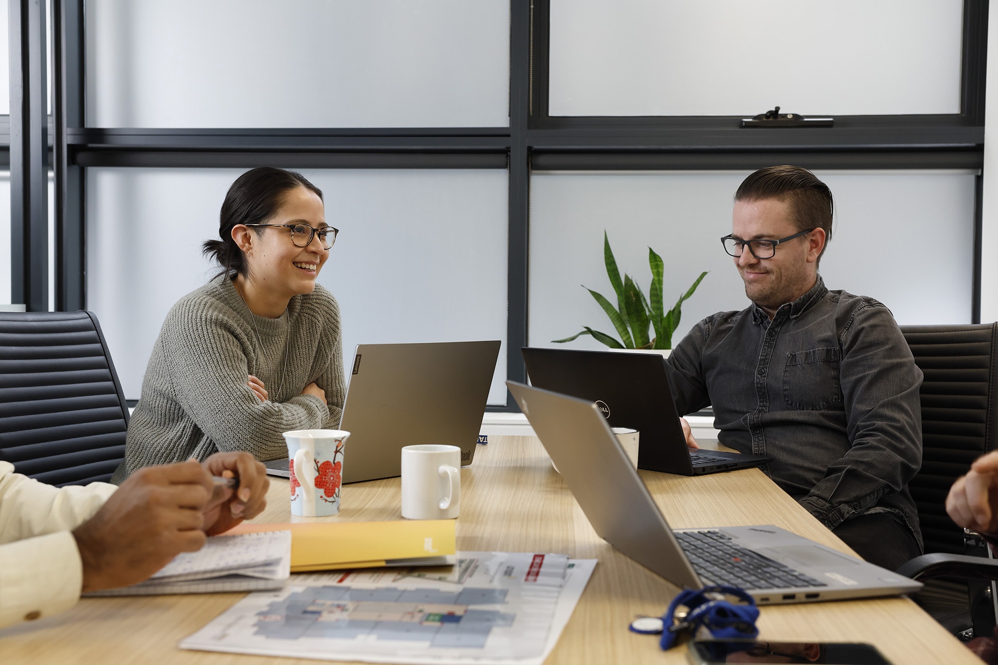 Four people sit around a conference table with laptops, papers, and coffee mugs. Two are smiling and engaged in conversation. There is a plant and large windows in the background.