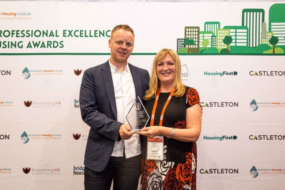 Two people stand smiling and holding an award together in front of a backdrop featuring logos and the text Professional Excellence in Housing Awards. The background includes stylized building graphics and sponsor names.