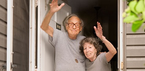 An elderly man and woman stand in a doorway, smiling and waving with one arm each. The man wears glasses and a gray shirt, and the woman is next to him, holding his waist. They appear happy and welcoming.