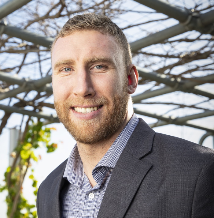 A smiling man with short light brown hair and a beard wearing a gray blazer and checkered shirt stands outdoors under a metal pergola with vines and bright sunlight.