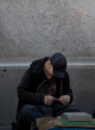 A person wearing a black jacket and cap sits on the ground against a gray wall, looking down at something in their hands. Personal belongings and a cardboard sign are placed in front of them.