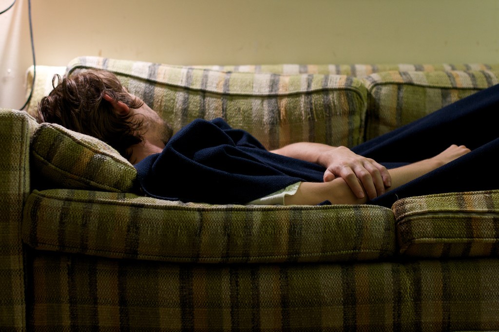 A person with brown hair is lying on a plaid-patterned couch, facing away from the camera, with their hands resting on their side and a dark blanket covering part of their body.
