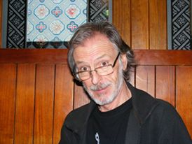 An older man with gray hair and a beard, wearing glasses and a black jacket, sits in front of a wooden paneled wall with patterned tiles above. He is looking at the camera and smiling slightly.