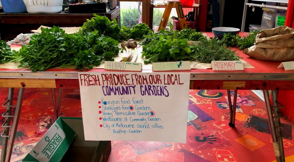 A table displays fresh herbs and greens labeled Pallard and Rosemary. A handwritten sign below reads Fresh Produce from Our Local Community Gardens with a list of garden contributors.