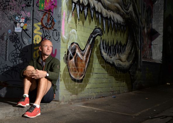 A man sits on the pavement in an alley covered with graffiti, including a large mural of a reptilian creature smoking a pipe. Sunlight highlights the man while the background remains mostly in shadow.