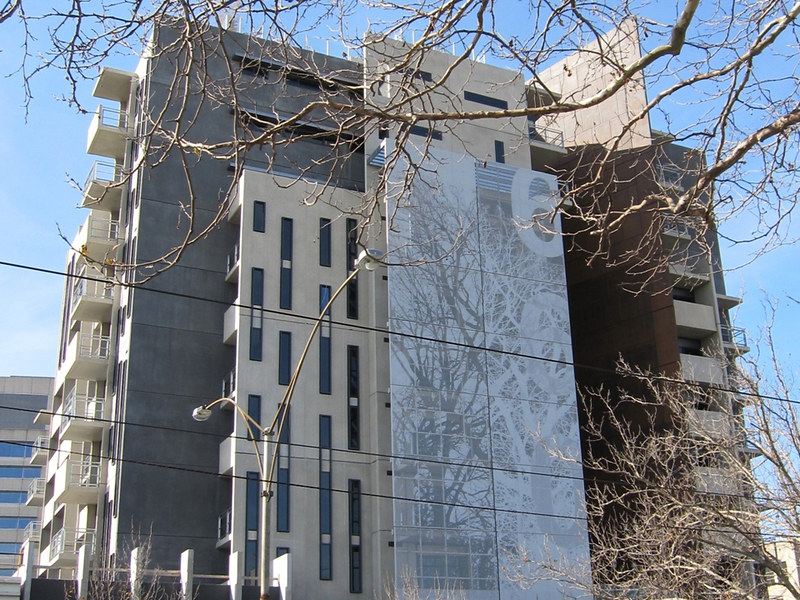 A modern, multi-story apartment building with a gray facade and vertical windows, partially obscured by bare tree branches, under a clear blue sky.