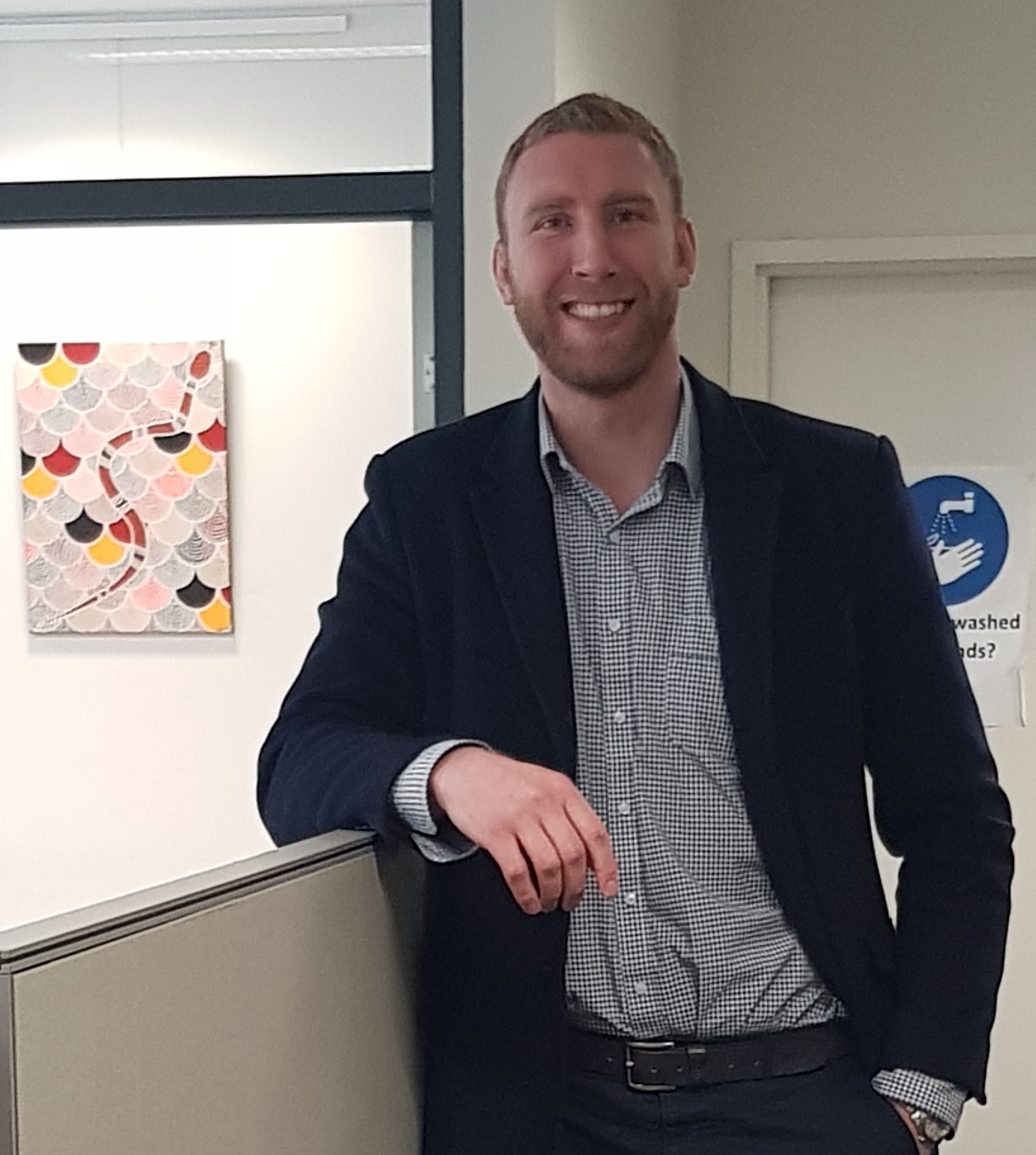 A man with short hair and a beard, wearing a dark blazer and checked shirt, smiles while leaning on an office partition. There is a colorful patterned artwork and a handwashing sign in the background.