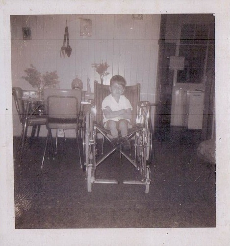 A young boy sits in a wheelchair in a room with a dining table and chairs. The photo is in black and white, giving it a vintage look. Flowers and utensils decorate the background wall.