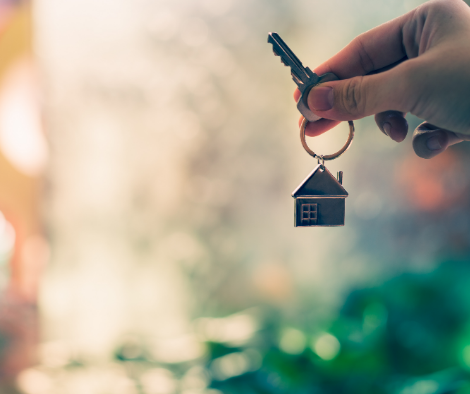 A hand holds a key with a house-shaped keychain in front of a blurred, colorful background, symbolizing homeownership or moving into a new home.