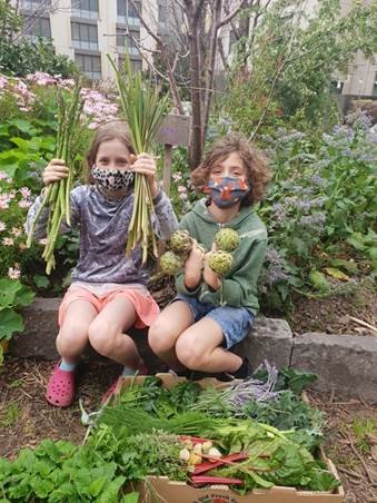 Two children wearing masks sit in a garden holding fresh vegetables, including asparagus and artichokes, with a box of assorted greens and herbs in front of them. Flowers and plants surround them.