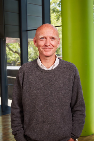 A bald man in a gray sweater stands indoors, smiling, with large windows, green pillar, and trees visible in the background.