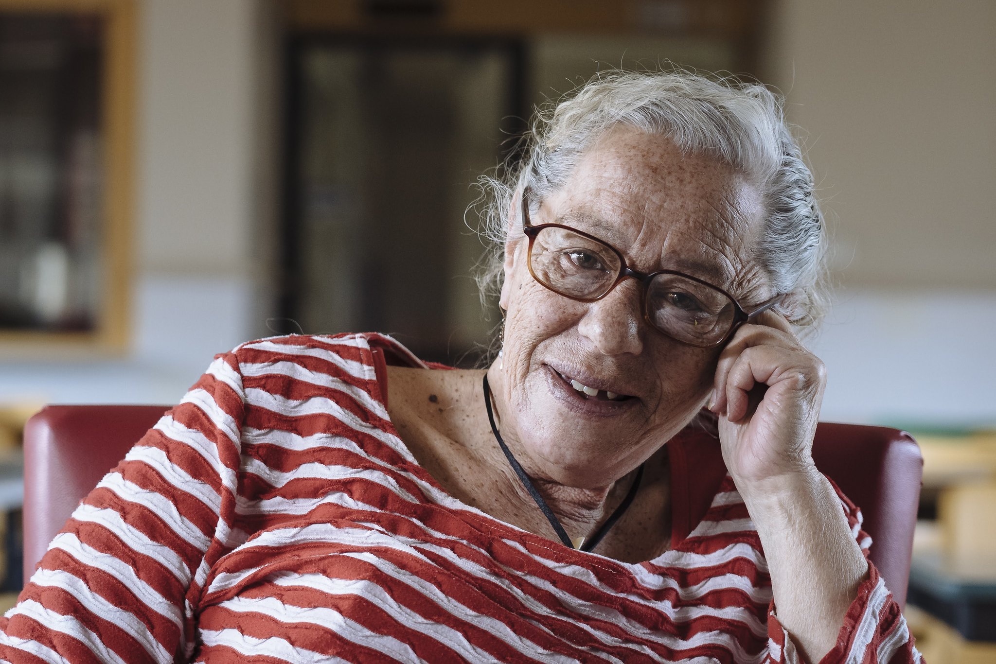 An elderly woman with gray hair and glasses smiles warmly, sitting in a chair and resting her head on her hand. She is wearing a red and white striped shirt, and the background is softly blurred indoors.