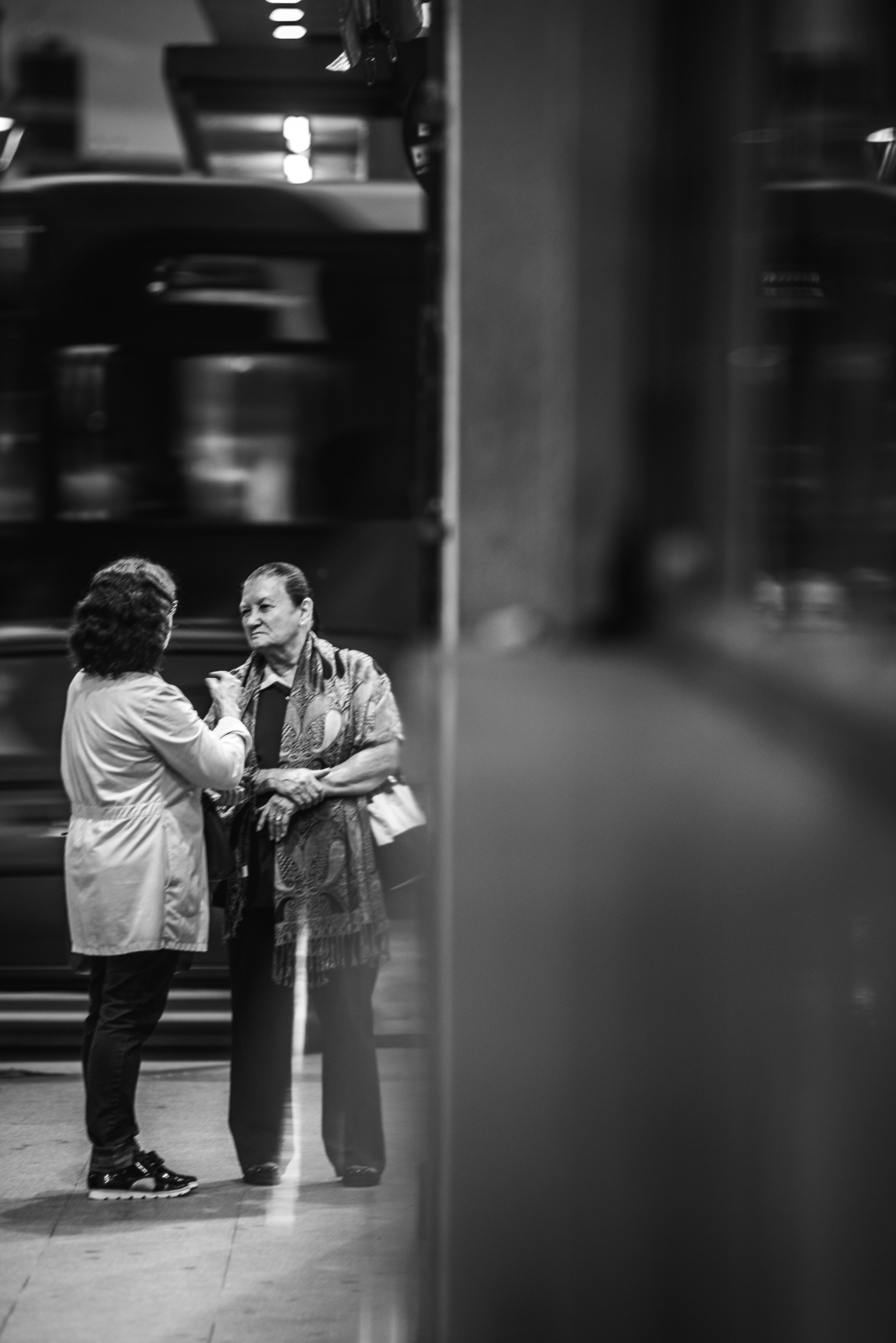 Two women stand and converse on a city sidewalk at night, with blurred lights and a moving vehicle in the background. The image is in black and white and has a reflective surface on the right side.