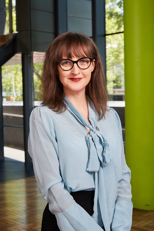 A woman with shoulder-length brown hair, wearing glasses, a light blue blouse with a bow, and a black skirt, stands indoors in front of large windows and a bright green column, smiling at the camera.