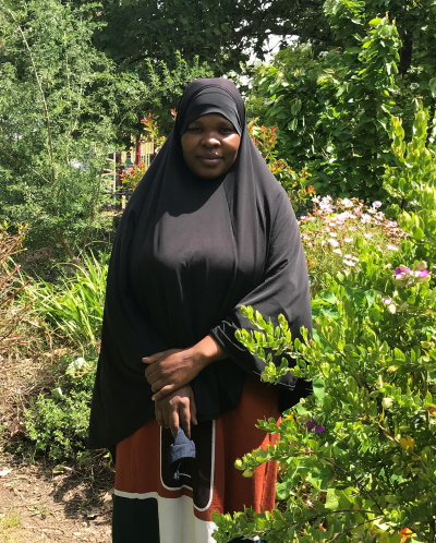 A woman wearing a black hijab and a long dress stands in a garden surrounded by green plants and blooming flowers, with sunlight filtering through the trees.