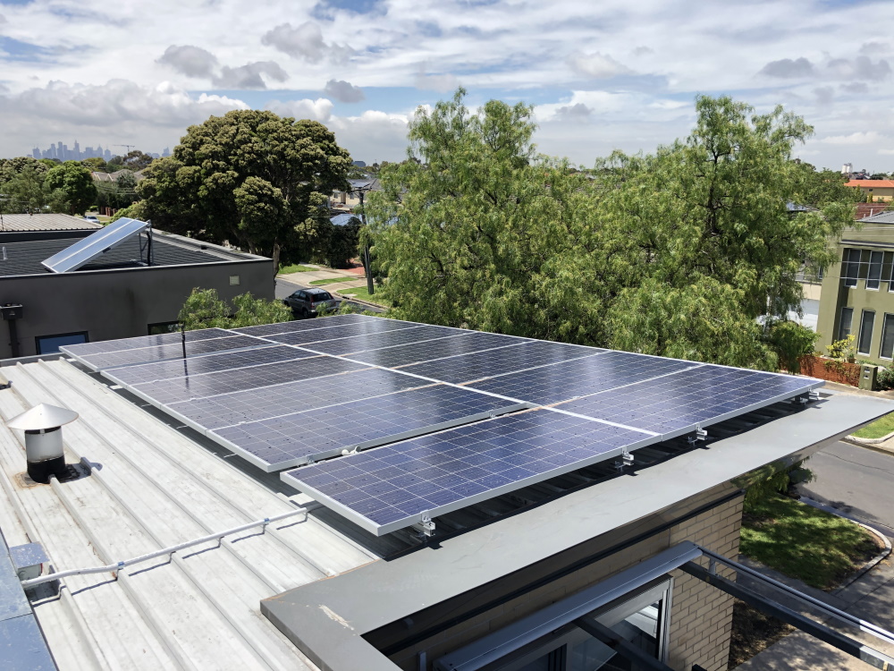 A set of solar panels installed on the flat roof of a building, surrounded by trees and other houses, under a partly cloudy sky. The city skyline is visible in the distant background.