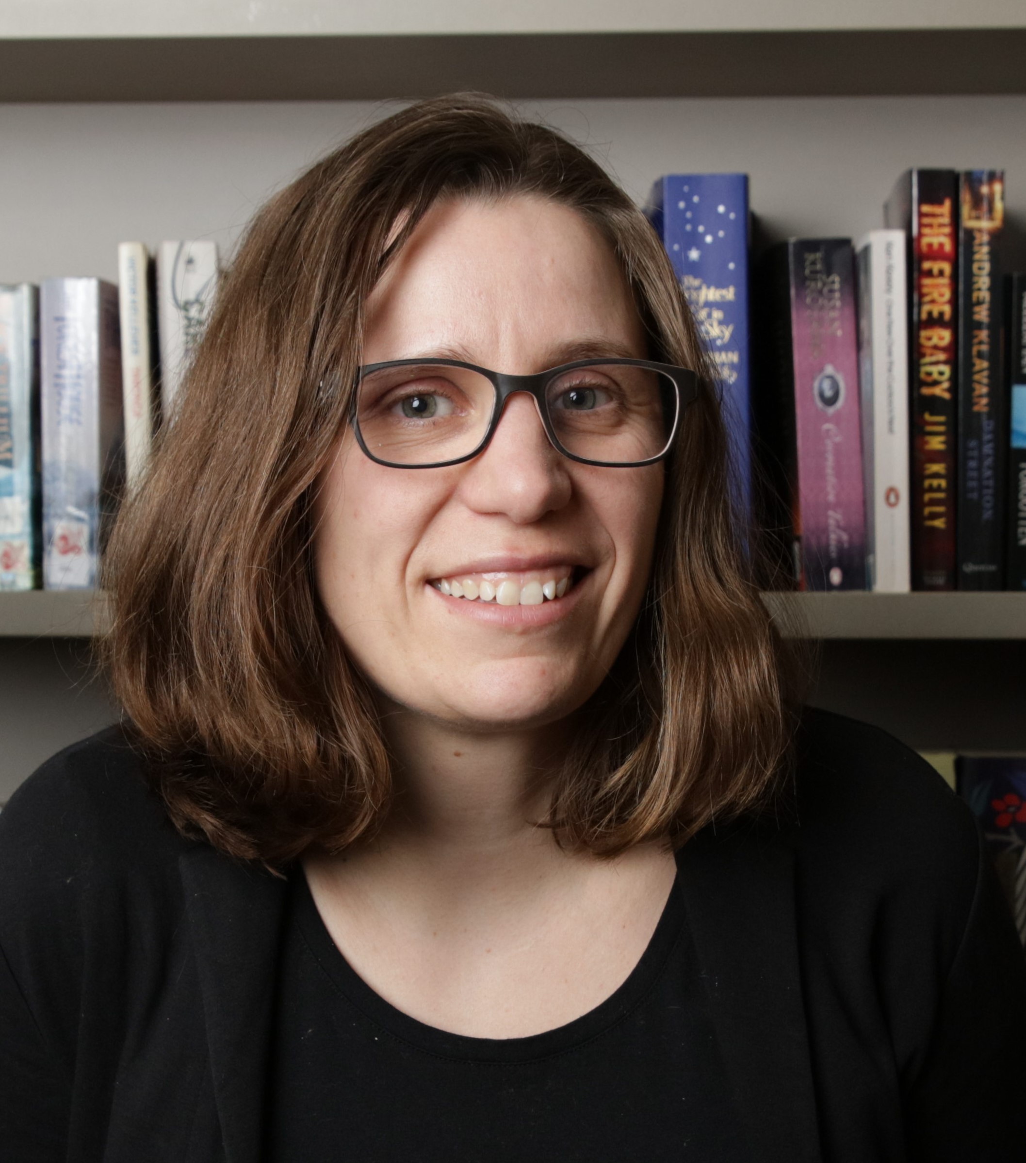 A woman with shoulder-length brown hair and glasses smiles in front of a bookshelf filled with books. She is wearing a black top and is positioned indoors.
