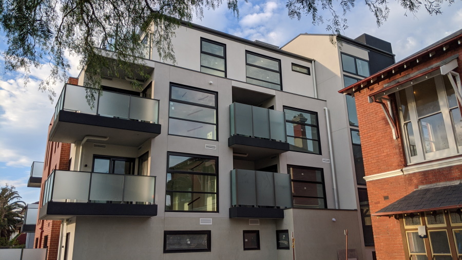 A modern three-story apartment building with large windows and glass balconies stands next to an older brick building under a partly cloudy sky. Tree branches are visible at the top left corner.