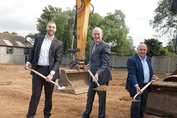 Three men in suits smile while holding shovels with dirt at a groundbreaking ceremony on a construction site. An excavator and fencing are visible in the background.