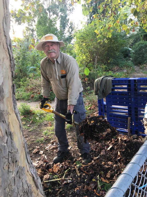 A man in a hat and work clothes smiles while shoveling soil or mulch in a garden area, surrounded by greenery and trees, with blue crates stacked nearby.