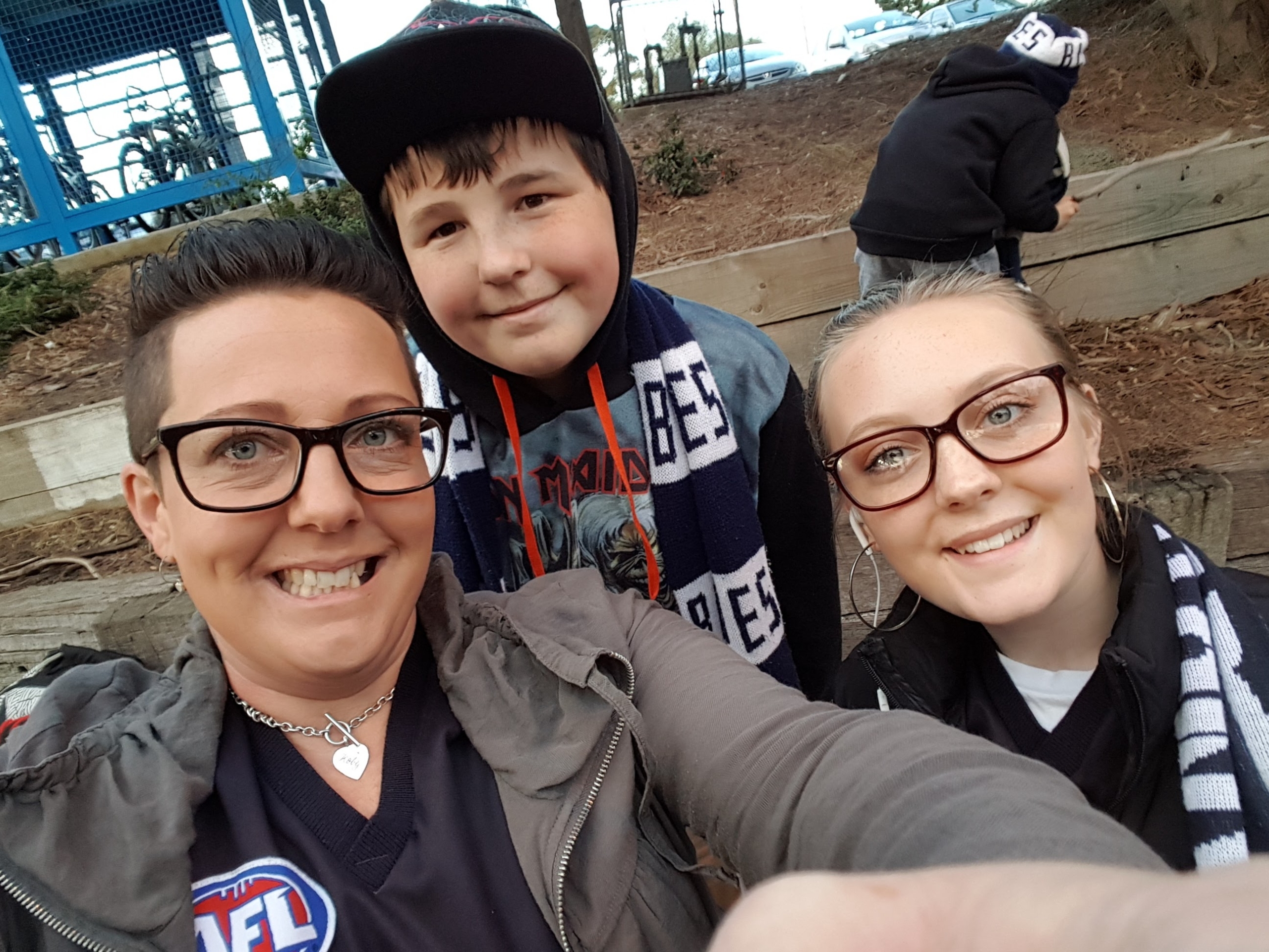 Three people, two women and a boy, smile for a selfie outdoors. They wear glasses and winter clothes, some with scarves and an AFL shirt. A child in a hoodie is in the background, near wooden steps and trees.