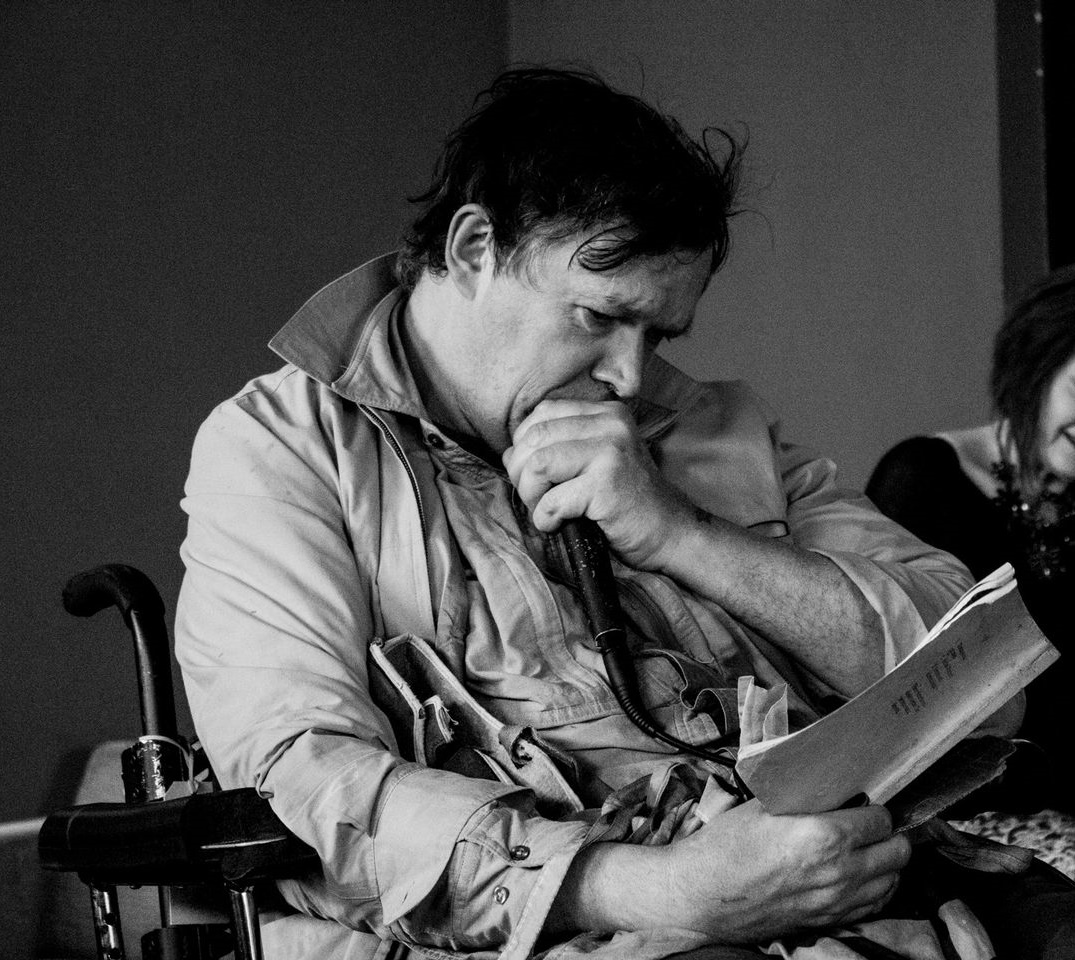 A man in a wheelchair, wearing a coat, sits with his head resting on his hand while intently reading a book or script. The mood appears contemplative. The image is in black and white.