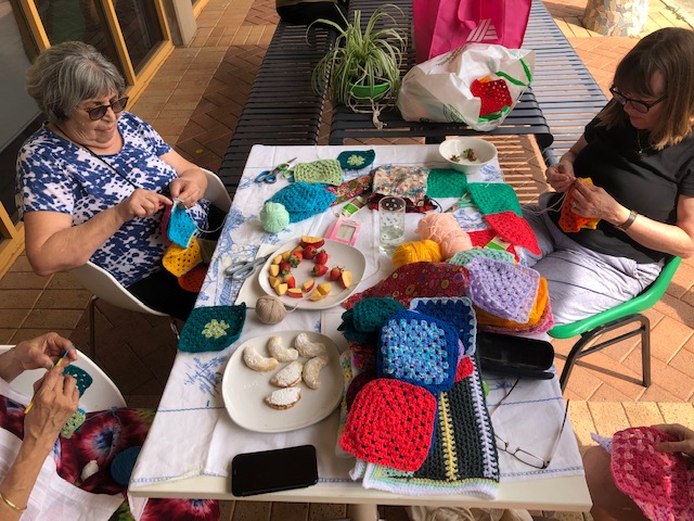 Three women sit around a table crocheting colorful granny squares. The table is covered with yarn, crochet projects, plates of snacks, and fruit. Sunlight streams in from nearby windows, creating a cozy, creative atmosphere.