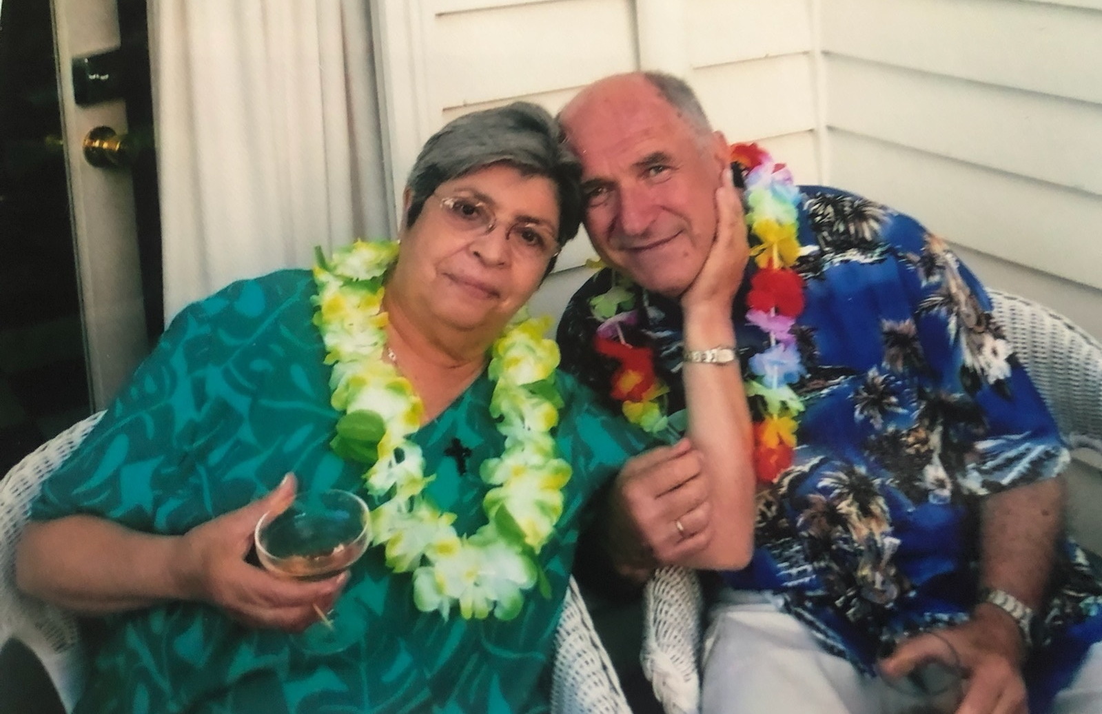 An older couple sits closely together on wicker chairs, both wearing colorful Hawaiian leis. The woman holds a drink and rests her hand on the mans face, while he smiles with his arm around her. They appear relaxed and happy outdoors.