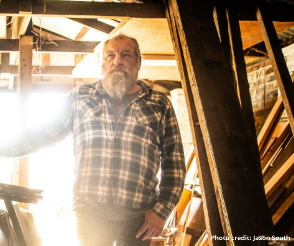 An older man with a grey beard wearing a plaid shirt stands in a sunlit, cluttered wooden workshop. Sunlight streams in from behind, creating a warm, rustic atmosphere. Photo credit: Jason South.