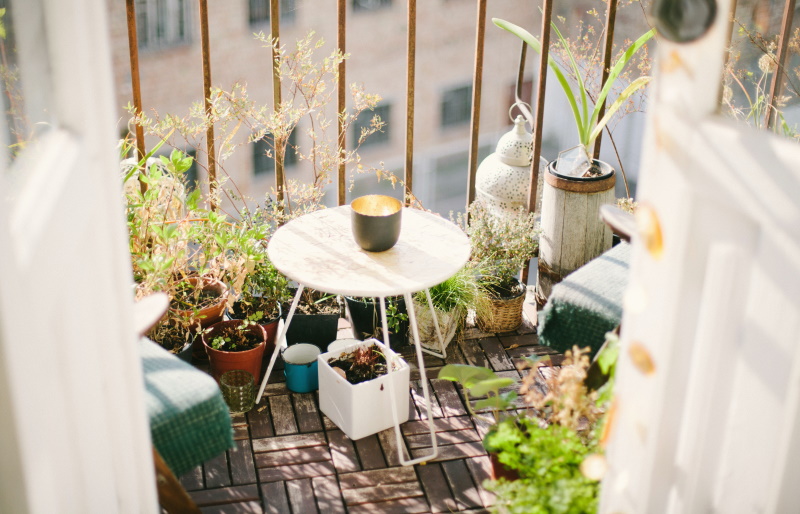 A small, sunlit balcony with a round table holding a cup, surrounded by potted plants and cushions. The scene is cozy and inviting, with soft natural light and a view of buildings beyond the railing.