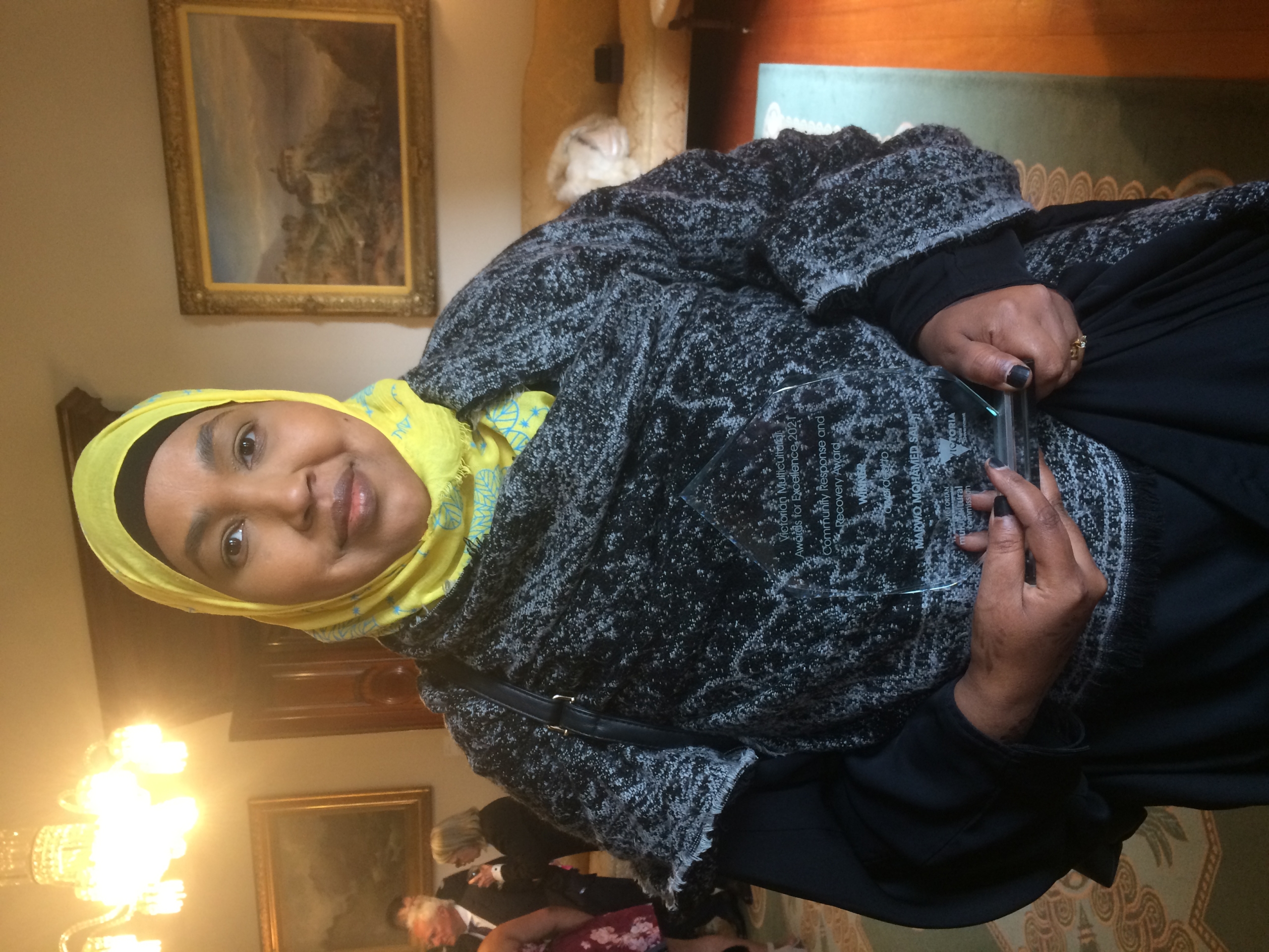 A woman in a yellow headscarf and patterned shawl holds a glass award, standing in a formal room with paintings and a chandelier. Other people are gathered in the background.
