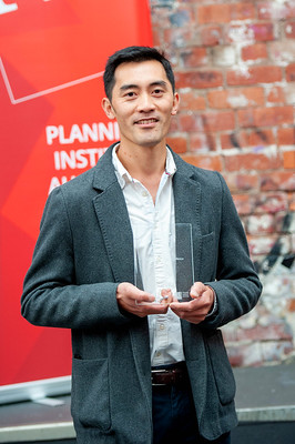 A man in a grey blazer and white shirt smiles while holding a clear rectangular award. He is standing in front of a red banner and a brick wall background.