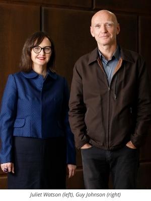 A woman in a blue jacket and glasses stands next to a man in a brown jacket and blue shirt. They are both facing the camera and standing against a dark wooden background.
