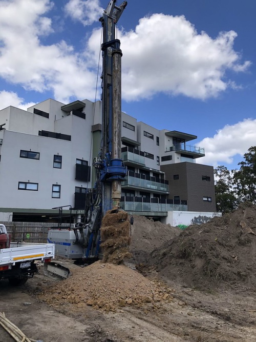 A drilling machine operates at a construction site near a modern apartment building, with soil and dirt piled around the equipment under a partly cloudy sky.