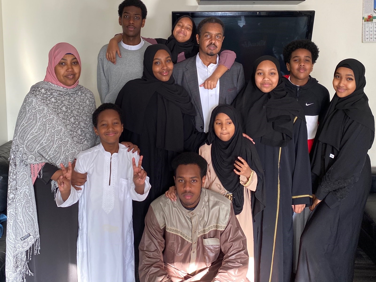 A family of twelve poses indoors, smiling at the camera. The group includes men, women, and children, many wearing traditional or modest clothing, with some women in hijabs. They stand closely together in front of a TV.