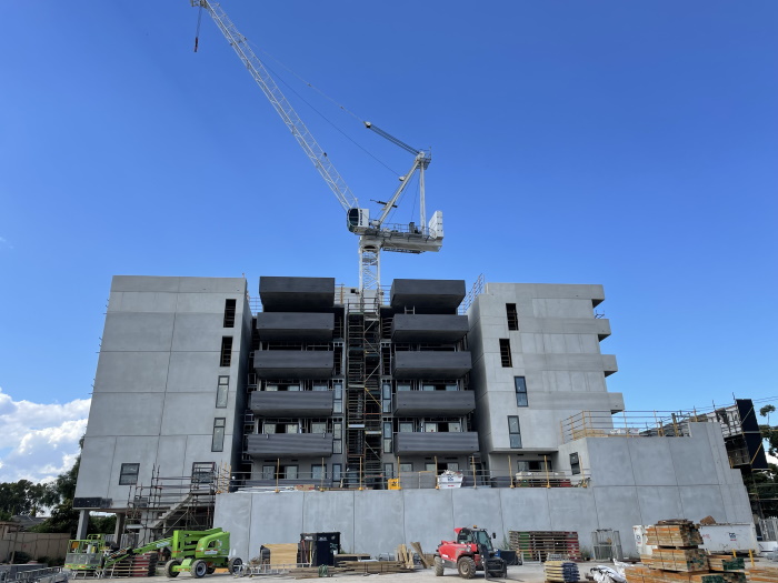 A large white crane towers over a multi-story concrete building under construction, with scaffolding and construction equipment visible in front of the structure against a clear blue sky.