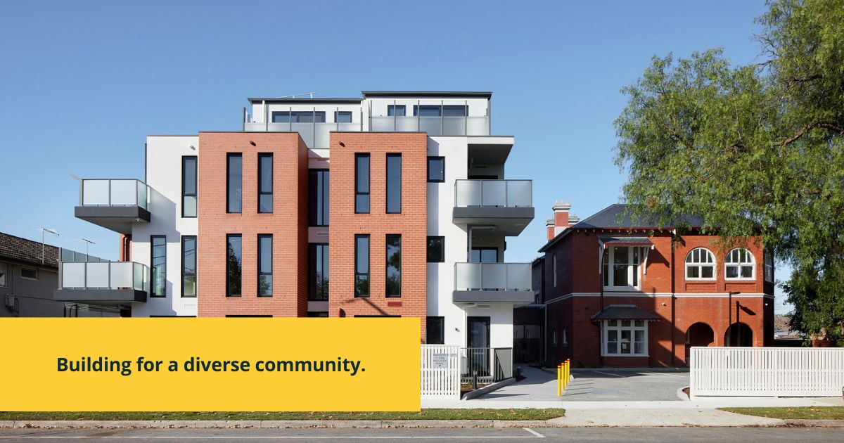 A modern apartment building with balconies stands beside a traditional red-brick house. In the foreground, a yellow banner reads, “Building for a diverse community.” Trees and a clear blue sky are visible.