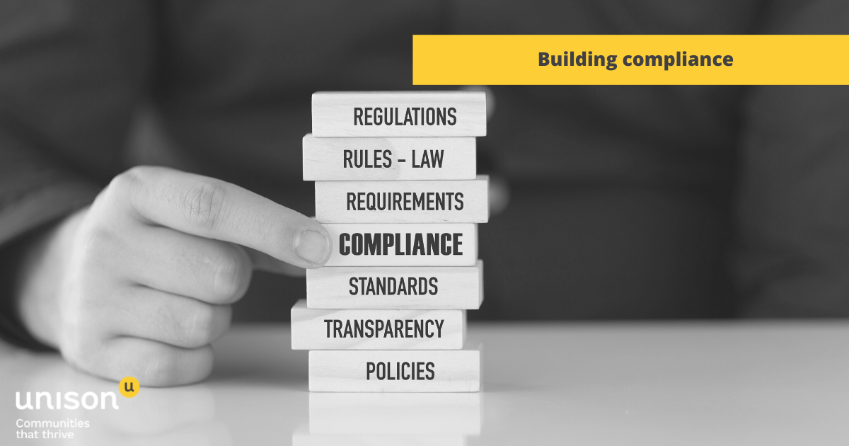 A person’s hand points to a wooden block labeled COMPLIANCE in a stack with other blocks labeled regulations, rules-law, requirements, standards, transparency, and policies. Text reads Building compliance. Unison logo is in the corner.