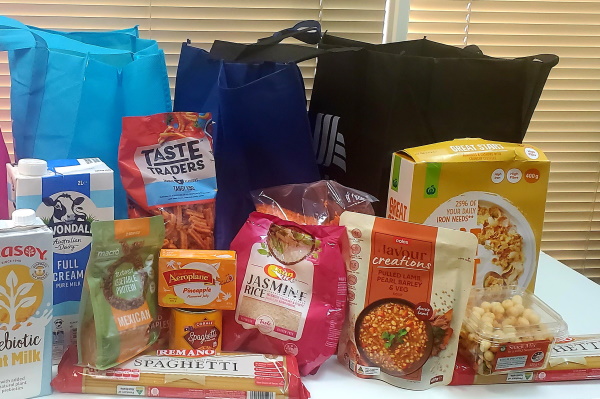 A variety of groceries, including pasta, rice, cereal, canned goods, and snacks, are displayed on a table in front of several reusable shopping bags.